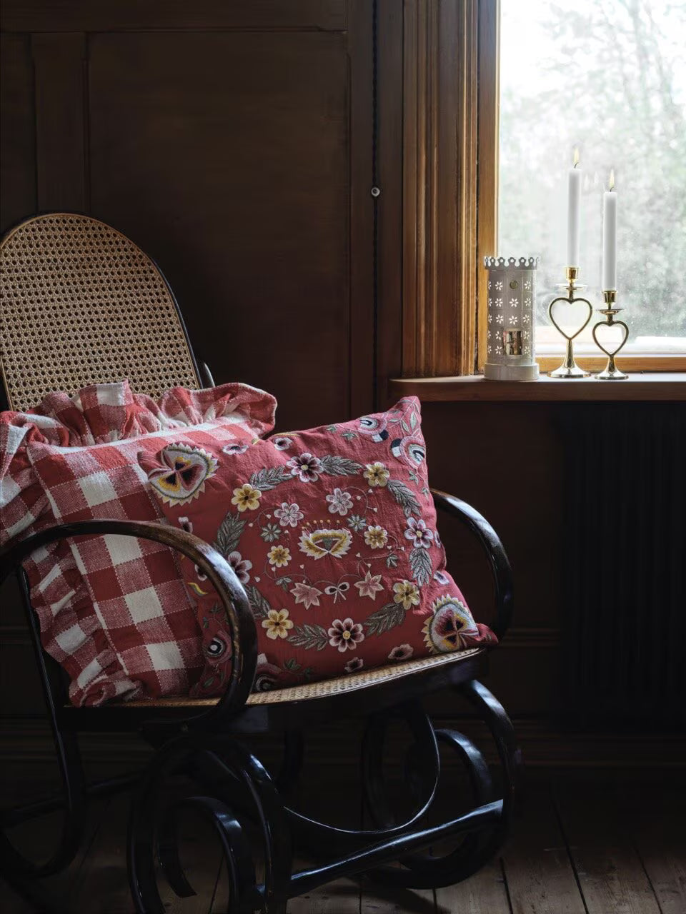 A wooden rocking chair with a cane backrest and a Cushion Cover Dalglans 45 x 45 cm from Strömshaga sits by a window adorned with two lit candles, a candle holder, and offers a view of trees outside.