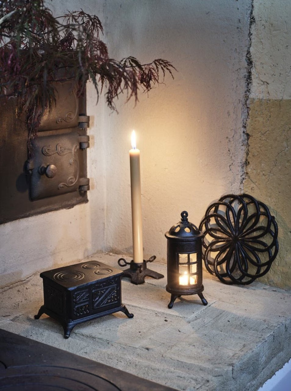 A lit Stove Candle Holder, Cast Iron by Strömshaga, sits on stone alongside a lantern, trivet, and vintage metal box in cozy home décor. Warm candlelight glows below a leafy branch, creating a peaceful atmosphere.