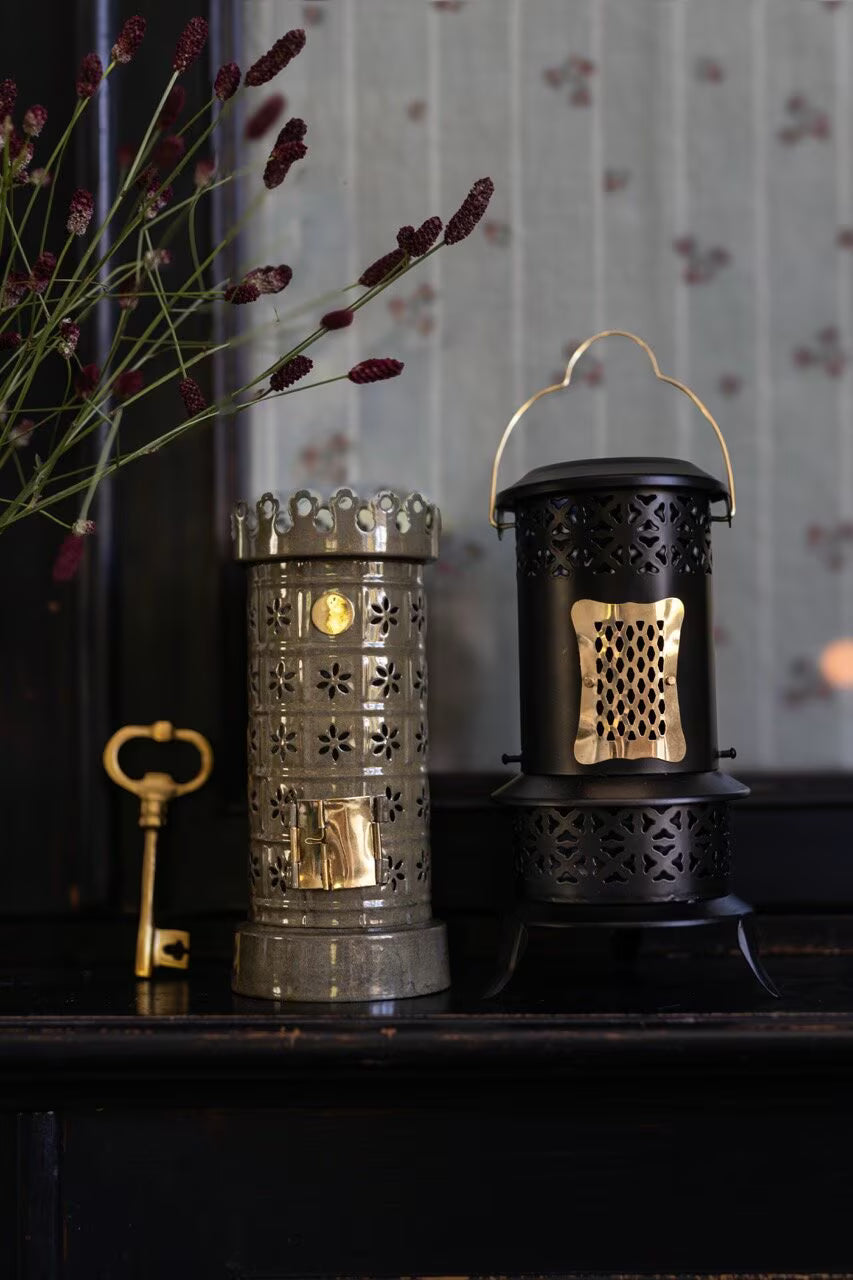 Two ornate lanterns—one silver, the other black with gold accents—rest on a dark wooden surface, creating a decorative display. Next to them lies the Strömshaga Bottle Opener Key in Antique Brass, its vintage design and subtle brass finish adding charm. Delicate flowers appear in the upper left corner, with a patterned curtain as the backdrop.