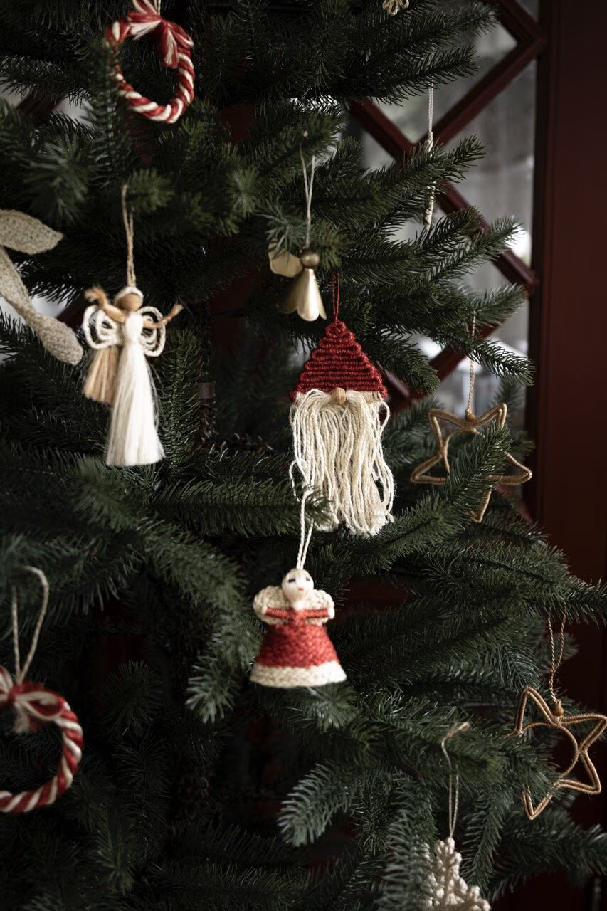 Handmade Christmas ornaments from Strömshaga, like the Nisse Santa Ornament crafted in jute with red and off-white accents, hang alongside an angel, stars, and candy canes on a green Christmas tree by a wooden-framed window—perfect for bringing cozy rustic charm to your holiday decor.