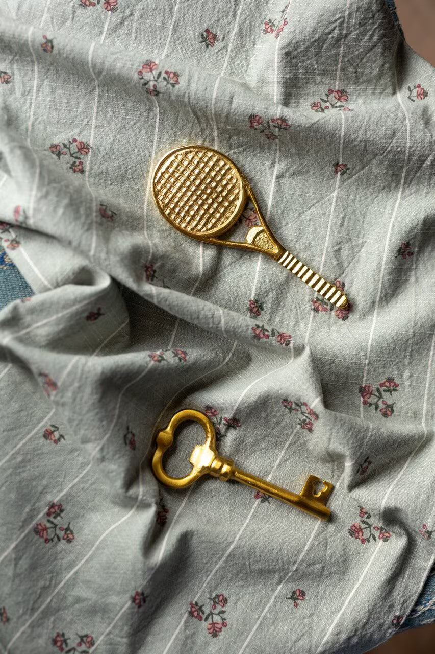 A gold bottle opener key with an antique brass finish from Strömshaga rests on a light blue, floral-patterned fabric with subtle stripes and small pink flowers, alongside a tennis racket-shaped object.