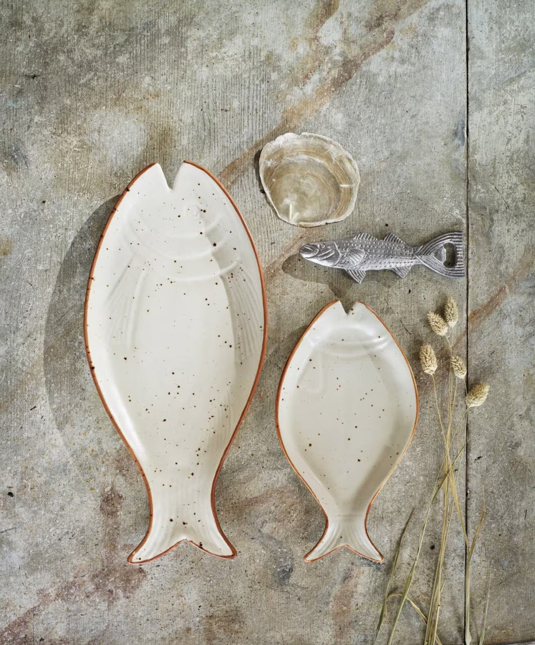 A large and a small fish-shaped ceramic plate are displayed on a textured surface with Strömshagas Bottle Opener Fish in Antique Silver, a small glass dish, and dried grasses.