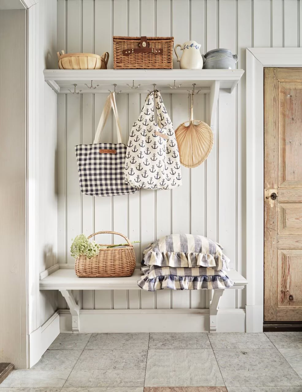 A white entryway with striped walls, a wooden bench topped with Strömshaga’s Cushion Cover Ida Stripe w. Frill in Blue & White (45x45 cm), pillows, baskets, and a wall shelf holding bags, pottery, and a wicker basket by a rustic wooden door.