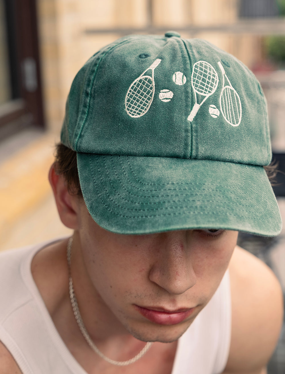 A person wearing the Fine Little Day Tennis Cap Embroidered in Cotton, Vintage Green, featuring tennis racket and ball graphics on the front, sitting outside.