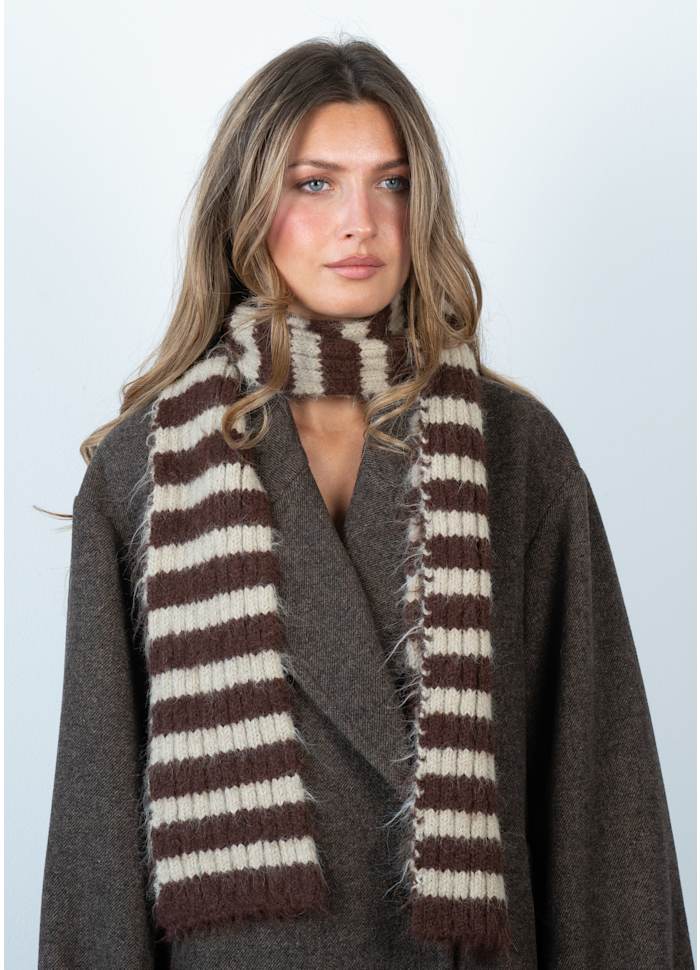 A woman with long, wavy hair wears a dark wool coat and a modern statement scarf—the Sui Ava Clara Slim Scarf in Brown and Beige—against a plain, light background.