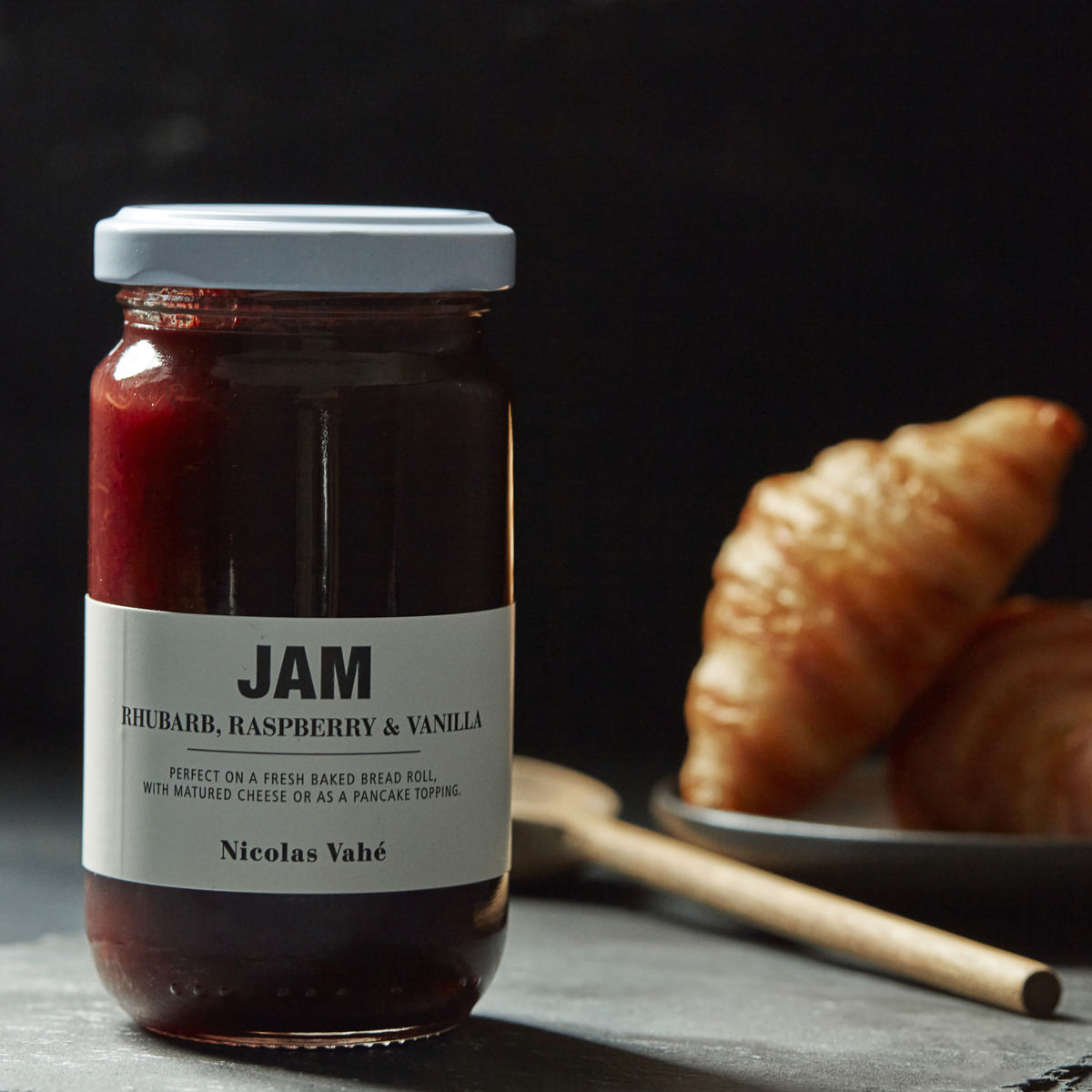 A jar of Nicolas Vahé Jam, Rhubarb, Raspberry & Vanilla rests on a dark surface beside a plate of croissants and a wooden spoon.