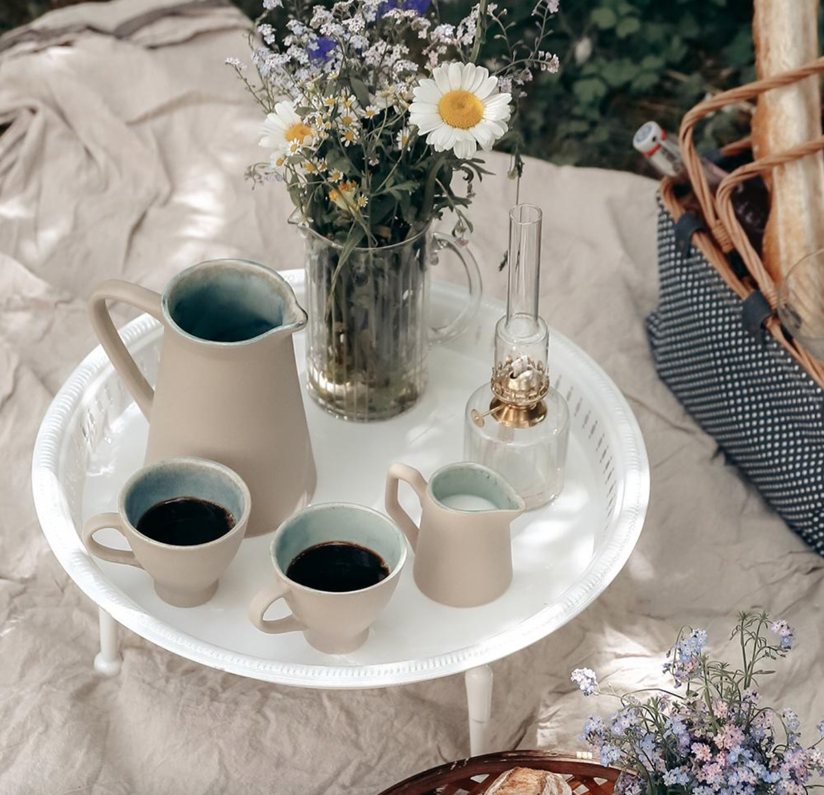 A round tray with cups, a jug, a small pitcher, and a glass vase of wildflowers sits on a blanket at a picnic, accompanied by the Elisabeth Oil Lamp in Clear Glass with Brass (Small, 25cm) from Strömshaga. Part of a basket and some bread are also visible.