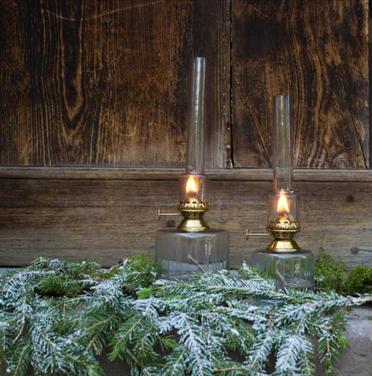 Two Strömshaga Elisabeth Oil Lamps in clear glass with brass burners (small, 25cm) cast a warm glow on a wooden surface, surrounded by snow-dusted pine branches against a rustic wooden wall.