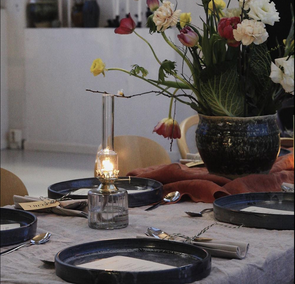 A dining table set with blue plates, rolled napkins, the Strömshaga Elisabeth Oil Lamp in Clear Glass with Brass (Small, 25cm), and a large floral arrangement with pink, white, and yellow flowers in a ceramic vase.