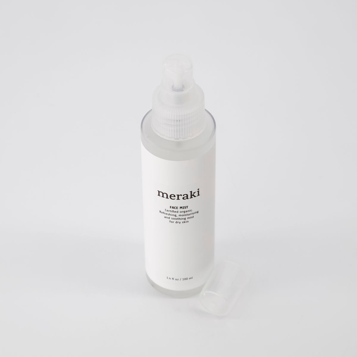 An upright, clear plastic spray bottle of Meraki Face Mist—Organic with Earth Marine Water—rests on a white surface, its translucent cap set beside it.