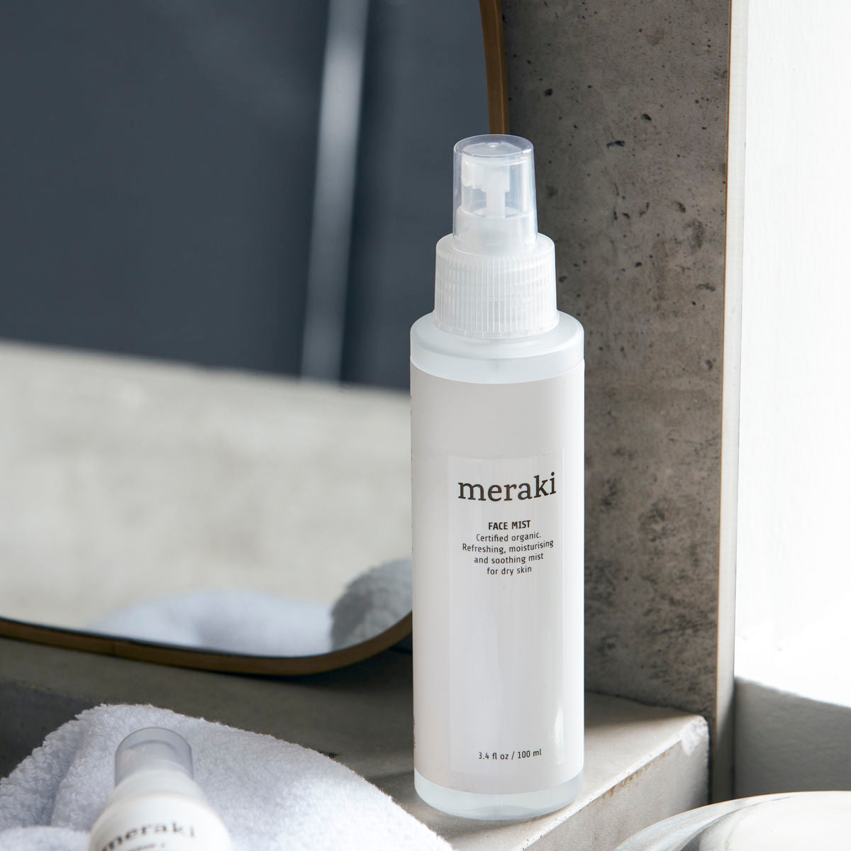 A white bottle of Meraki Face Mist, Organic with Earth Marine Water, rests on a bathroom counter near a mirror, accompanied by a towel and another partially visible bottle—ideal for achieving a radiant complexion.