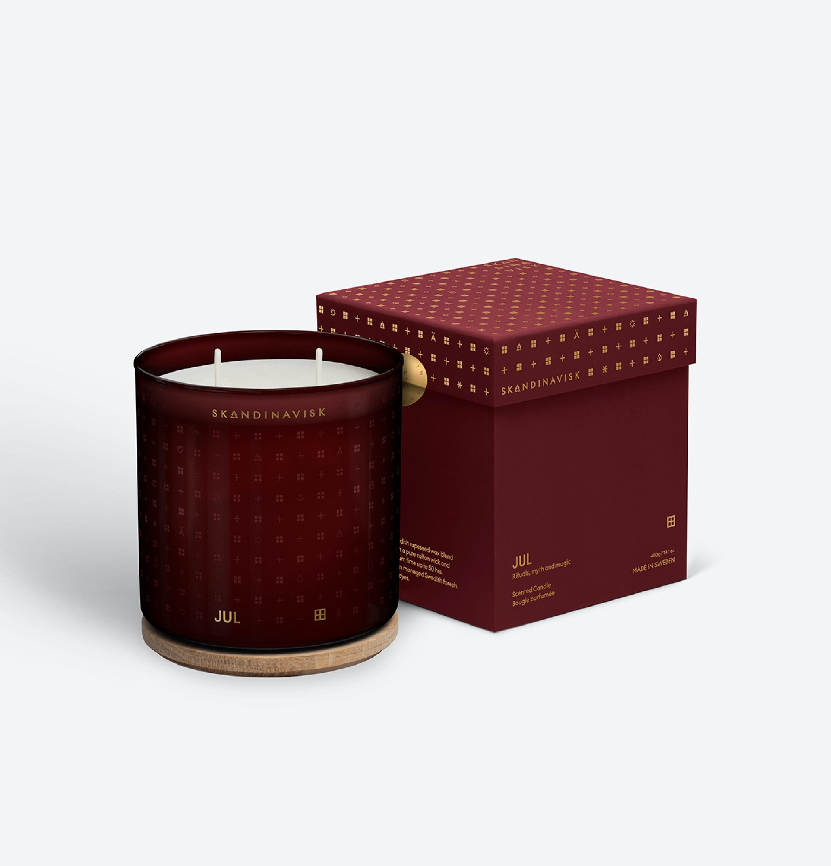 The Skandinavisk Seasonal Jul Scented Candle, 2 Wick, 400g, with Swedish rapeseed wax and a wooden base, sits beside a matching red box adorned with gold text and patterns against a light background.