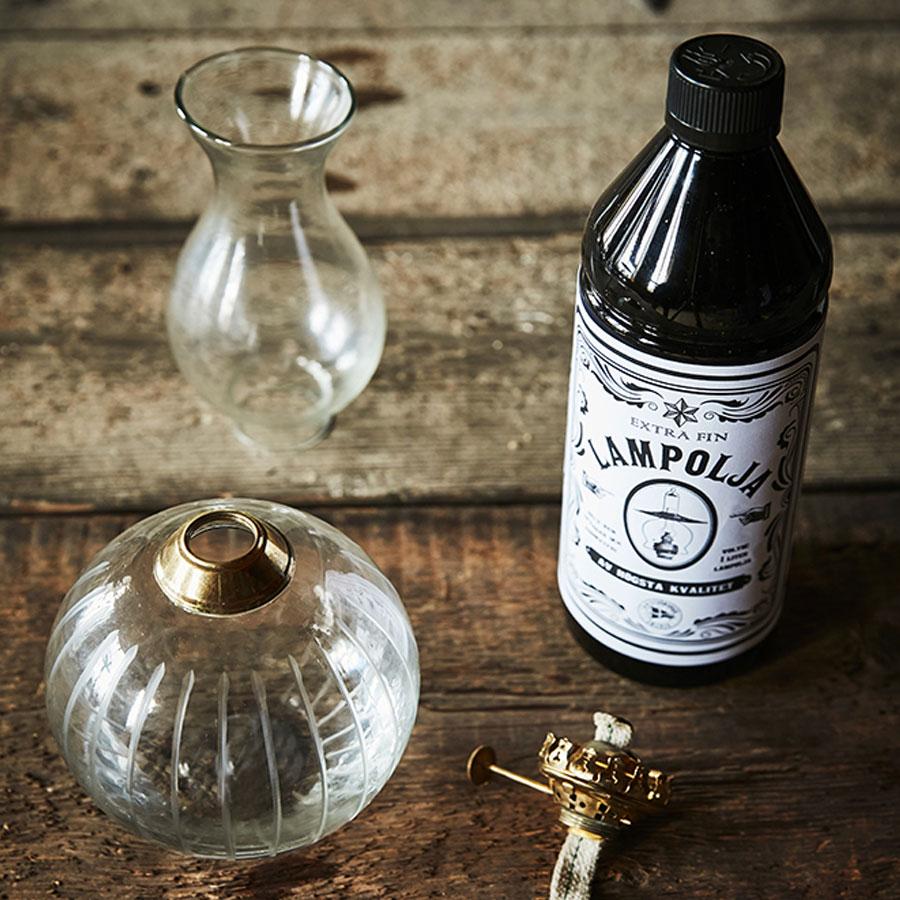 A clear glass lamp, chimney, brass burner top, and Strömshaga Lamp Oil 1 Liter bottle are arranged on wood, showcasing the importance of high quality lamp oil for kerosene lamps.