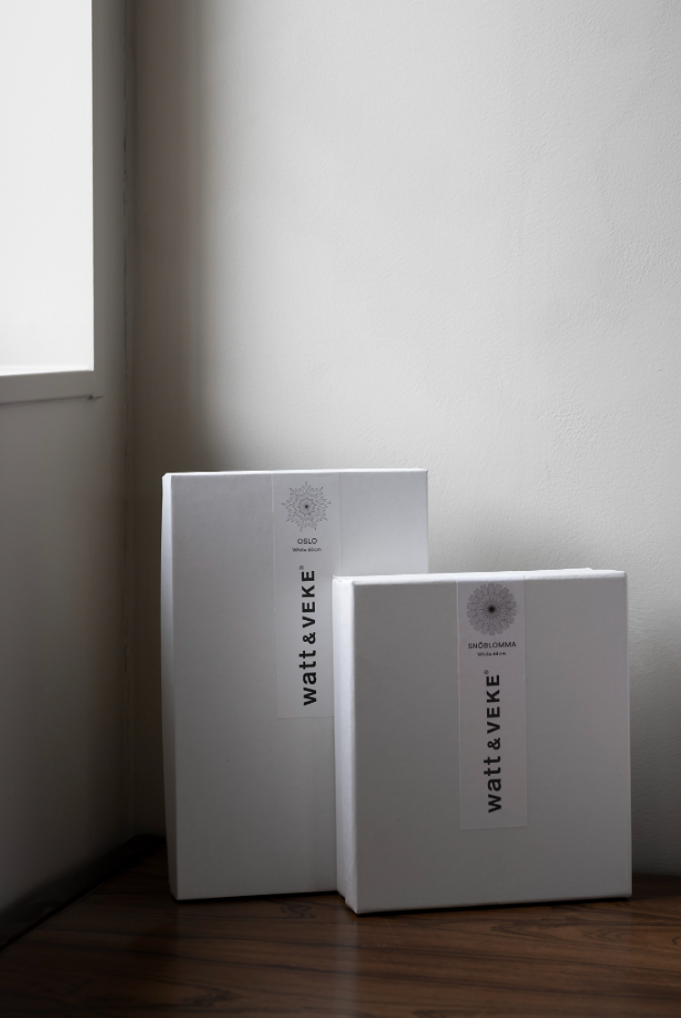 Two white rectangular boxes labeled Watt & Veke, containing the Star Eldig in Paper in White (52 cm), sit on a wooden floor in a bright room with white walls and sunlight coming through the left window.
