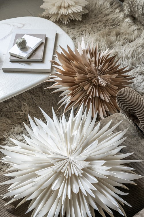 The Watt & Veke Star Maja in Paper, Gold (60cm) adds a decorative starburst charm to the cozy scene, displayed on a plush beige rug beside a coffee table and brown corduroy chair.