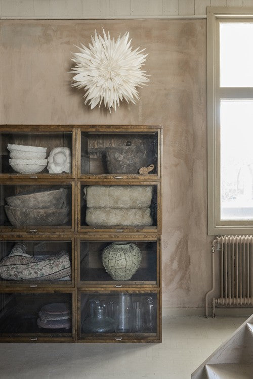 A wooden cabinet with glass-fronted shelves holds bowls and jars. Above, the Watt & Veke Star Maja in Paper, White 60cm, adds charm. A window and radiator are on the right, while a white finish brings elegance to the space.