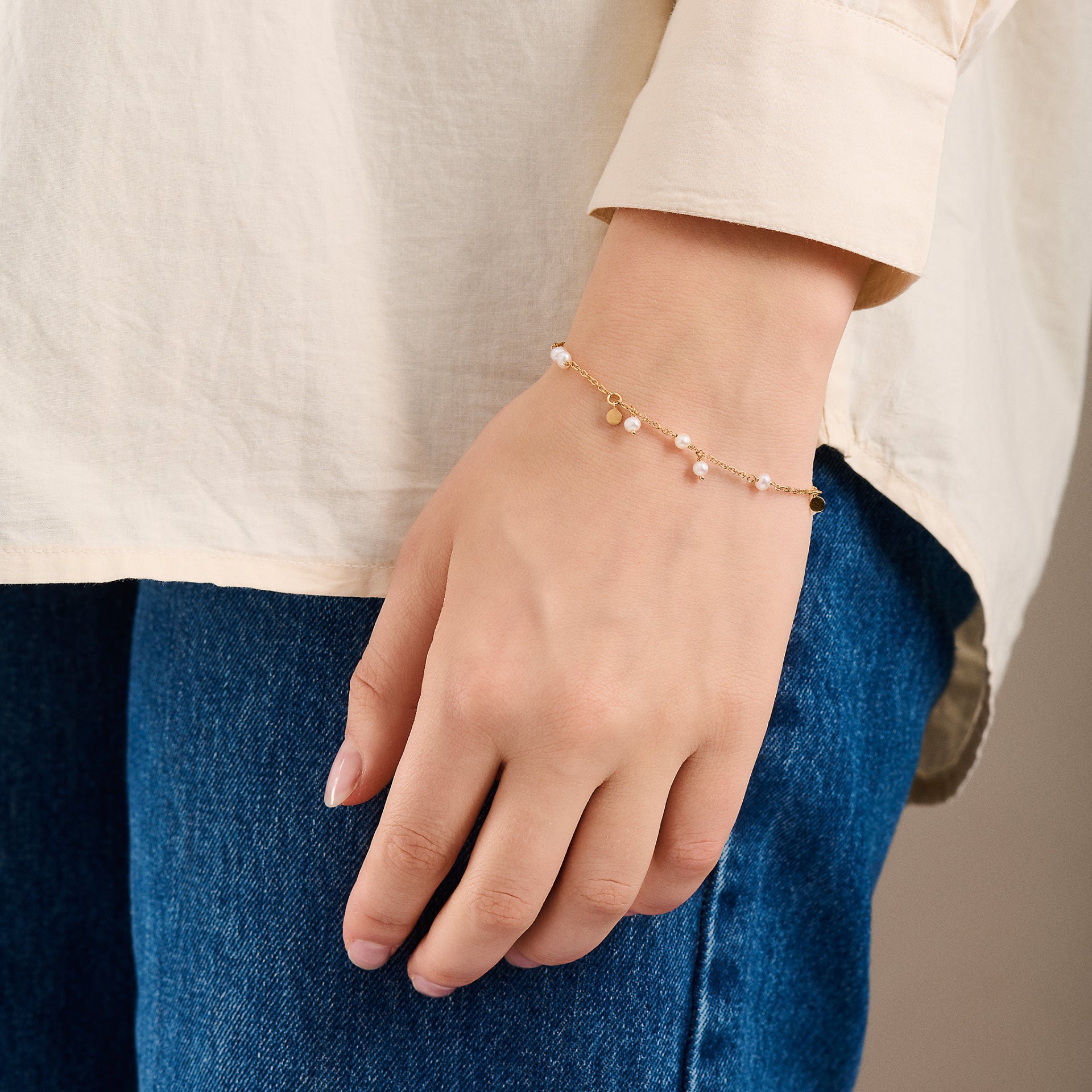 A person in a cream long-sleeve shirt and blue jeans showcases the Pernille Corydon Ocean Pearl Bracelet in Gold, featuring delicate beads, charms, and pearl pendants inspired by nature.