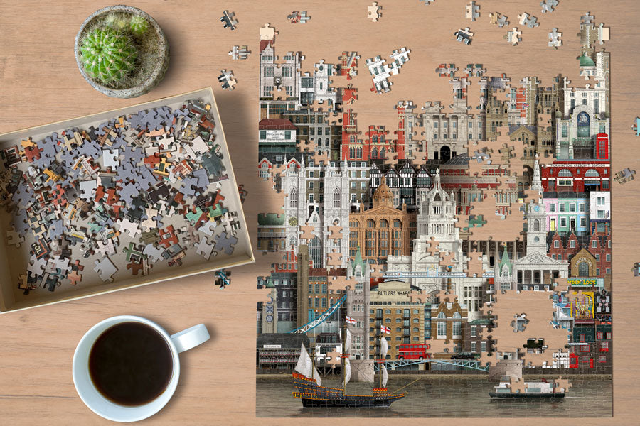 The Martin Schwartz Puzzle London City, 1000 Pieces, is partially completed on a wooden table next to its box of loose pieces, a cup of coffee, and a small potted cactus.