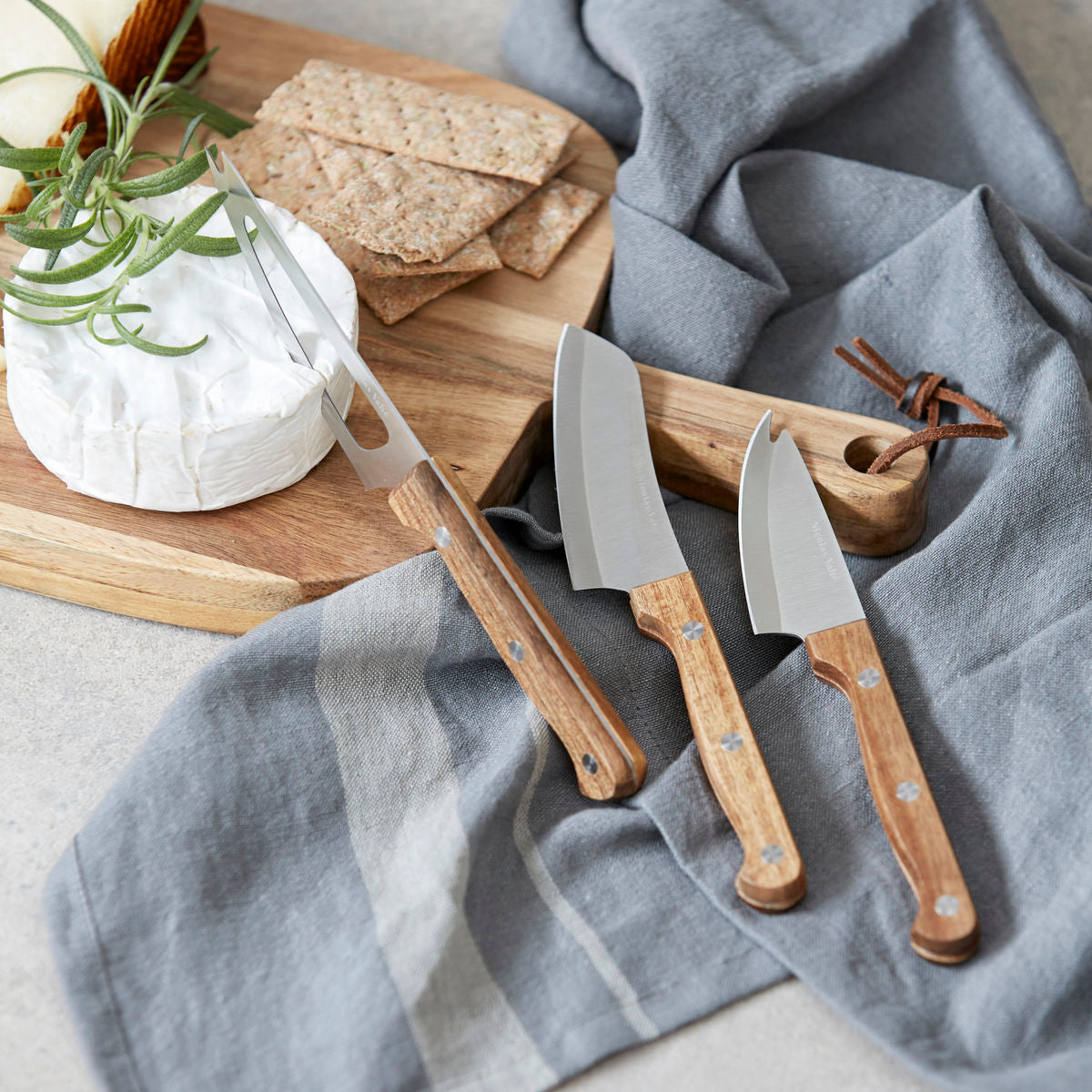 The Nicolas Vahé Cheese Knives Set of 3, featuring acacia wood handles, rests on a gray cloth beside a cheese wheel, crackers, and rosemary sprig on a wooden board. Comes in a gift box.