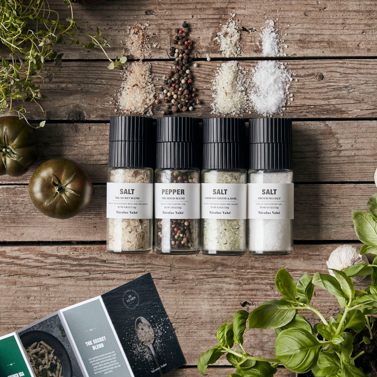 Four Nicolas Vahé spice grinders from the Gift Box, Favourite Collection—featuring French Sea Salt, Parmesan Cheese & Basil Salt, The Secret Blend Salt, and The Mixed Pepper Blend—each with a ceramic grinder for effortless use, are arranged on a rustic wooden table surrounded by herbs, tomatoes, peppercorns, and spilled salt. Partially visible printed cards in the lower left corner hint at gourmet blends perfect for elevating everyday meals.