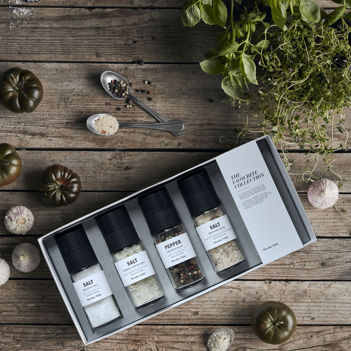 A beautifully arranged Nicolas Vahé Gift Box, Favourite Collection featuring four gourmet ceramic grinders—French Sea Salt, Parmesan Cheese & Basil Salt, The Secret Blend Salt, and The Mixed Pepper Blend—rests on a rustic wooden table surrounded by fresh herbs, tomatoes, garlic cloves, and a spoonful of mixed spices.