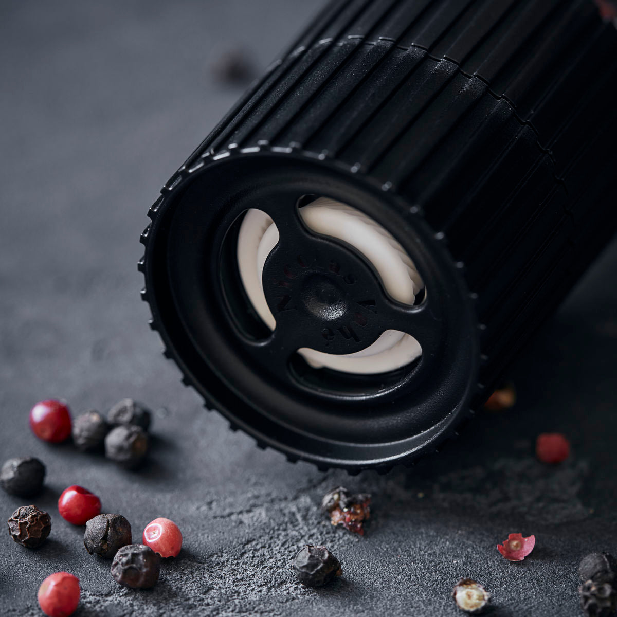 Close-up of the Nicolas Vahe Pepper Mix Grinder with black & pink peppercorns, featuring a ceramic grinder. It lies on its side on a dark surface, surrounded by colorful pepper mix and scattered whole peppercorns.