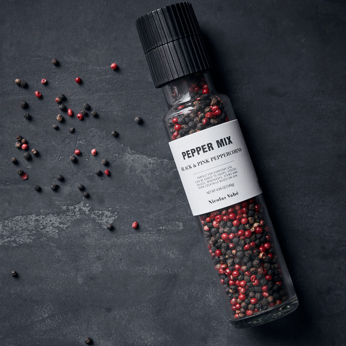 A clear Nicolas Vahe Pepper Mix Grinder with black & pink peppercorns sits on a dark gray surface, surrounded by scattered peppercorns. The label reads: Pepper Mix Grinder w. Black & Pink peppercorns.