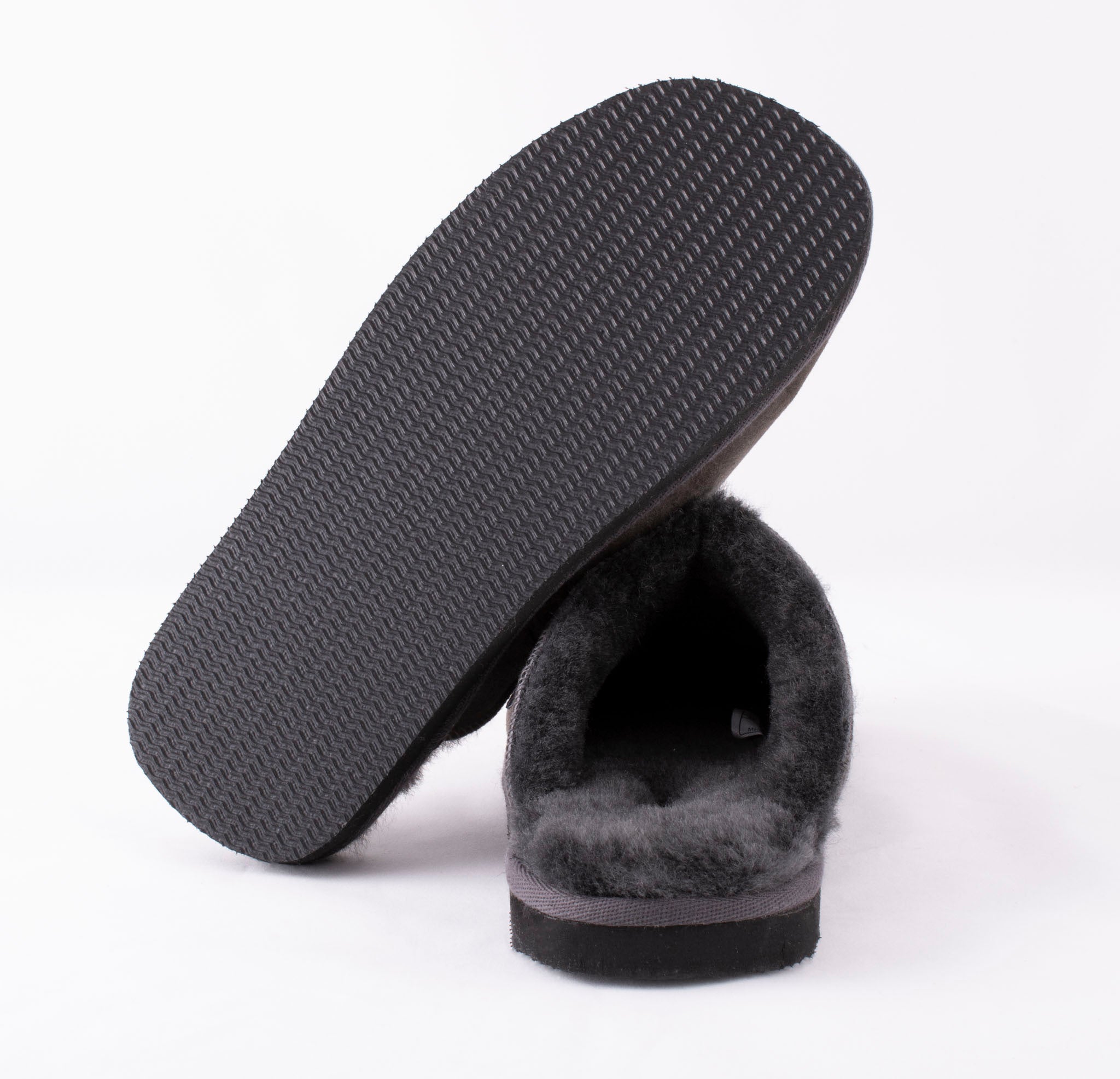 A pair of Shepherd of Sweden Hugo men's sheepskin slippers in asphalt grey; one stands upright to show its textured rubber sole, while the other highlights the soft, fluffy slip-in design, both set against a white background.