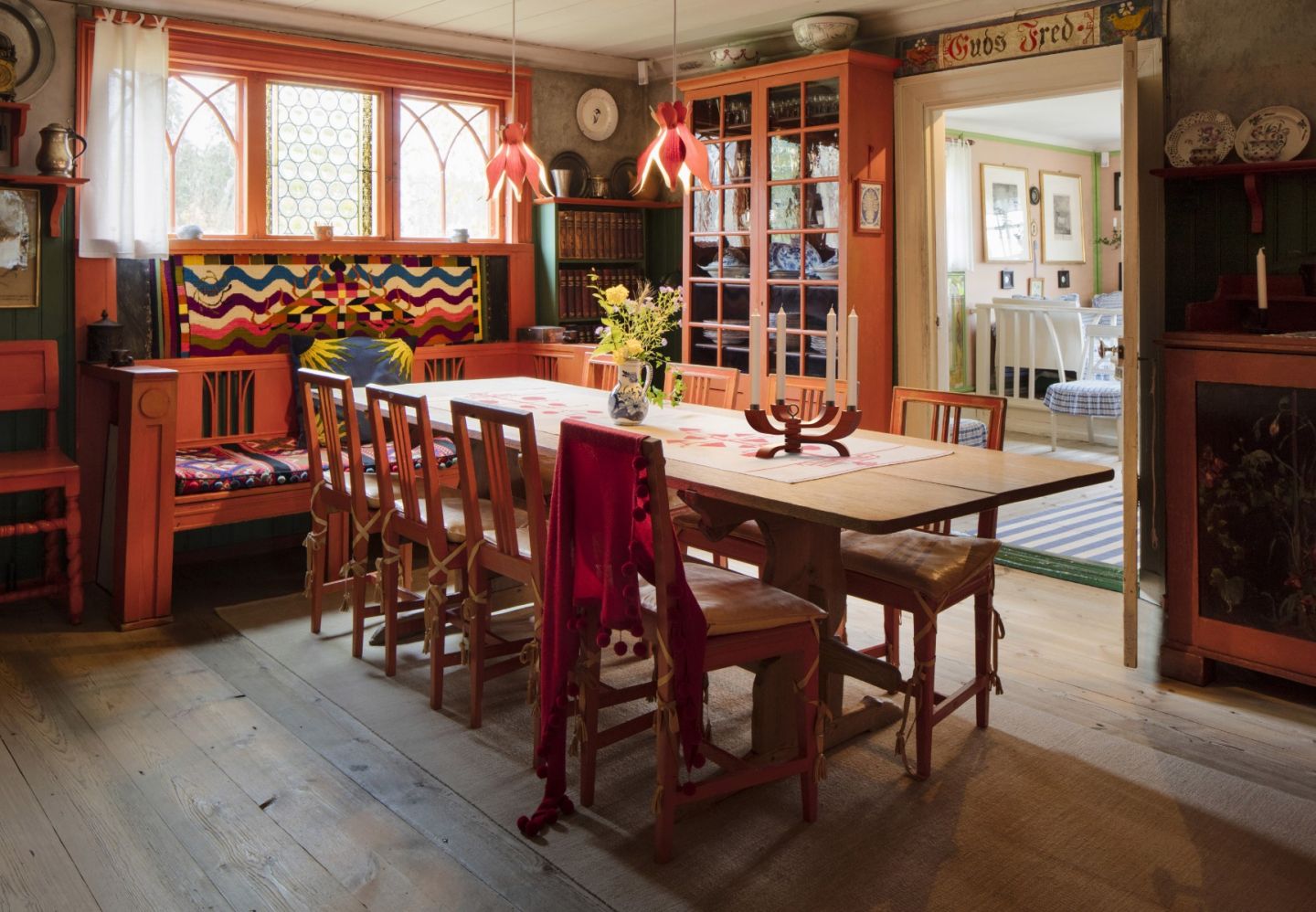 A cozy dining room with wooden floors and red furniture, including a long table and chairs. Colorful textiles, flowers, and the Design House Stockholm Nordic Light Candle Holder 4 Arms in Red enhance the warm, welcoming atmosphere. Sunlight streams in from an adjoining room.