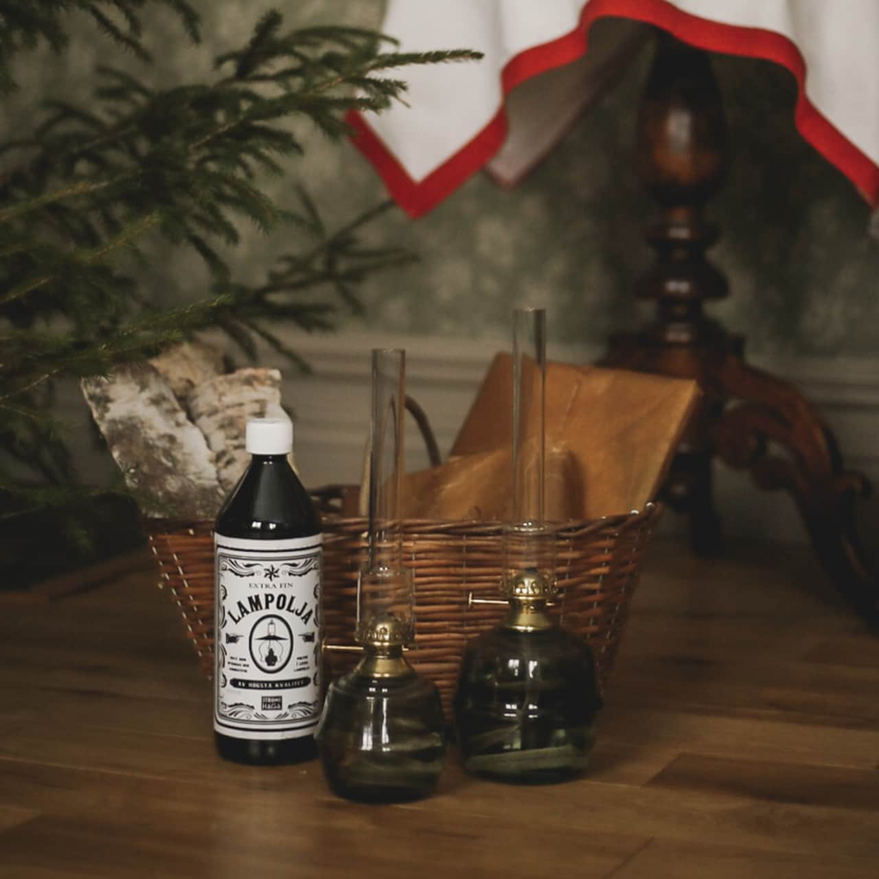 A wicker basket with firewood, a bottle of Strömshaga Lamp Oil 1 Liter, and two high-quality kerosene lamps sit on a wooden floor near a table with a white and red cloth.