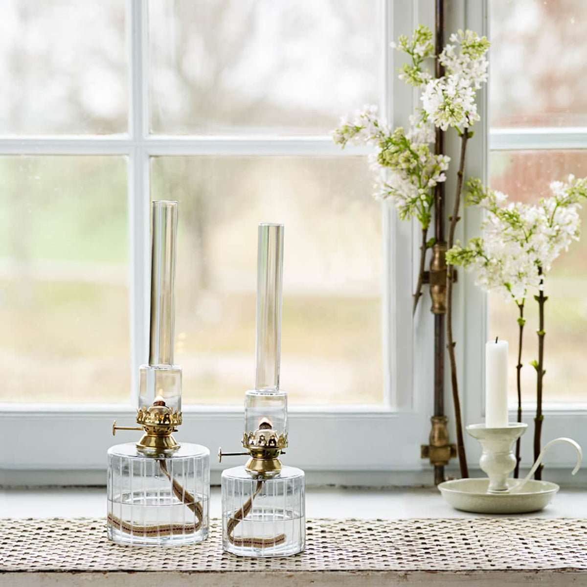 Two Strömshaga Elisabeth Oil Lamps in clear glass with brass burners (small, 25cm) sit on a woven mat in front of a window. Next to them are white flowers arranged in a vase and a single white candle on a green holder, creating a cozy, rustic atmosphere.