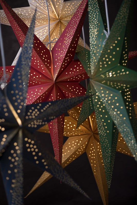 The Watt & Veke Star Skina Christmas White/Silver 80 cm lantern shines warmly among other perforated stars in red, gold, green, and white, their decorative holes glowing softly against a dark background.