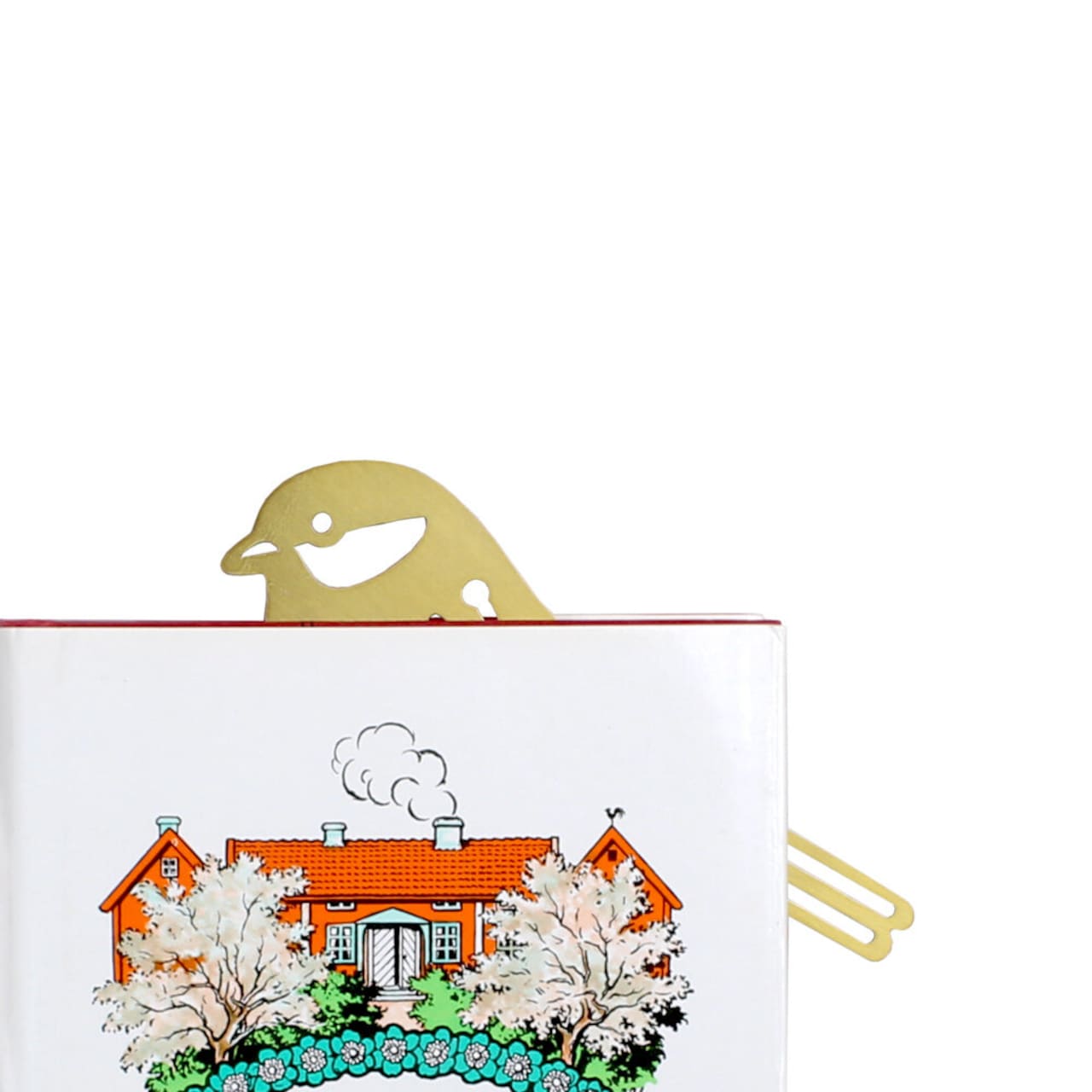 The Strömshaga Book Marker Bird in Brass peeks from a closed book with an illustrated cover featuring a red house, white flowering trees, and green bushes.